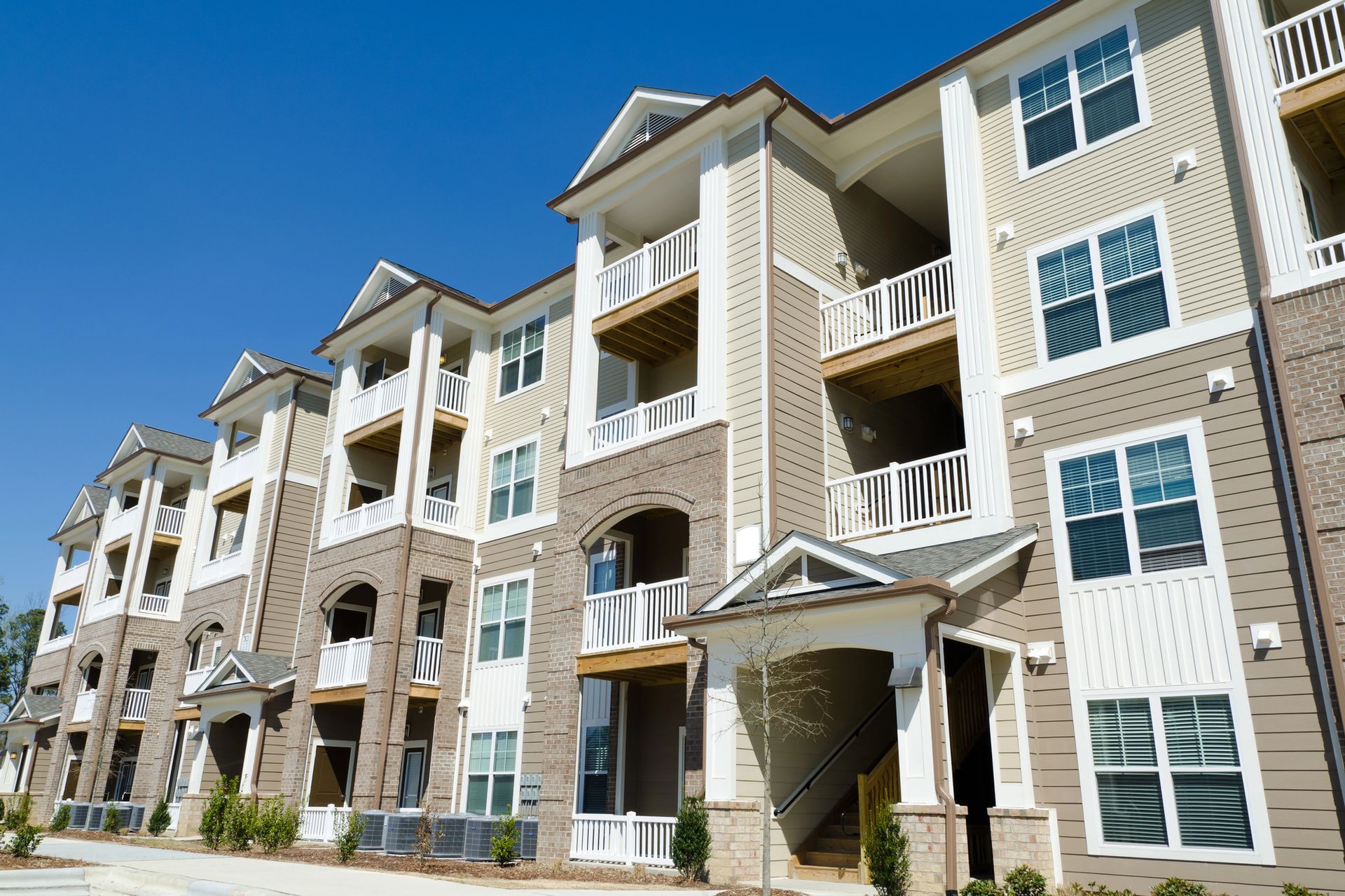 A multi-story apartment building with beige siding, brick accents, and balconies under a clear blue sky.