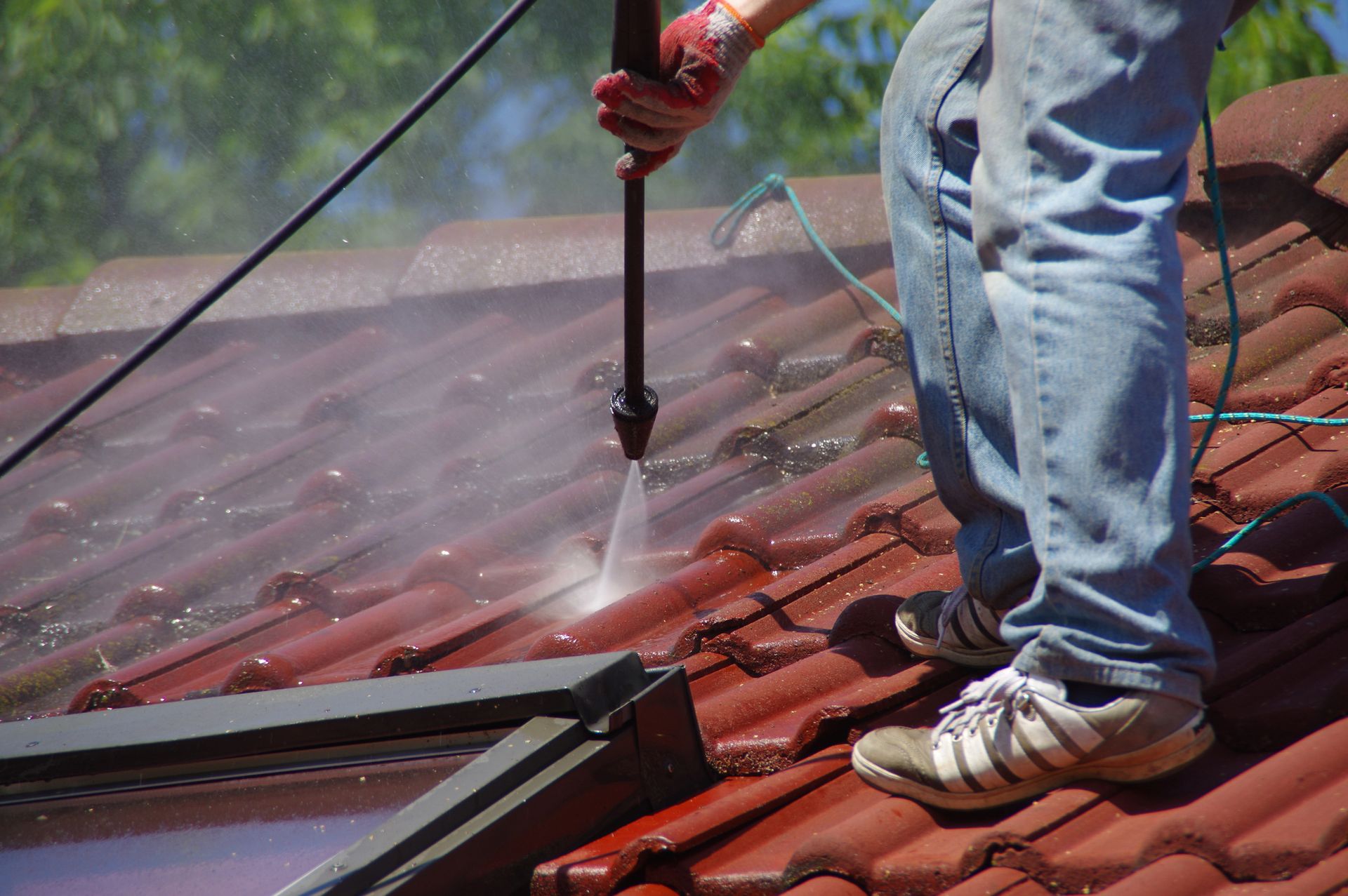 A person wearing jeans and sneakers uses a pressure washer to clean red roof tiles.