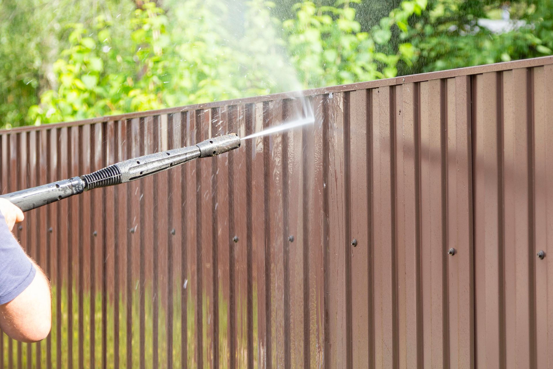 A person uses a pressure washer to clean a brown, corrugated metal fence.