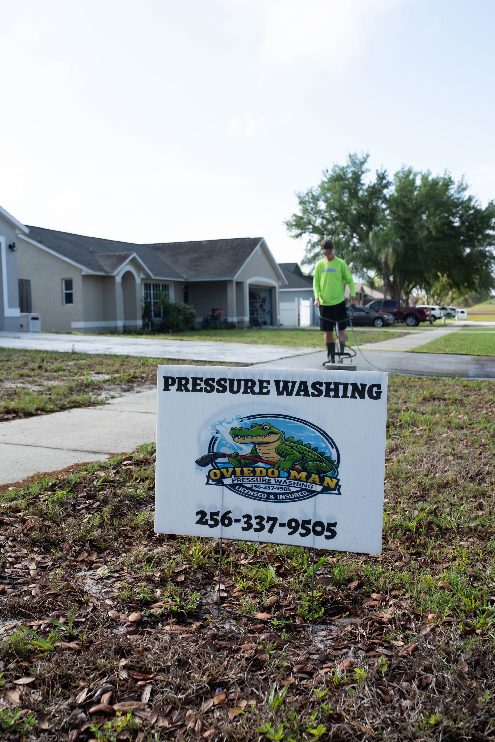A pressure washing sign in a grassy yard featuring an alligator logo and phone number, with a person working nearby.