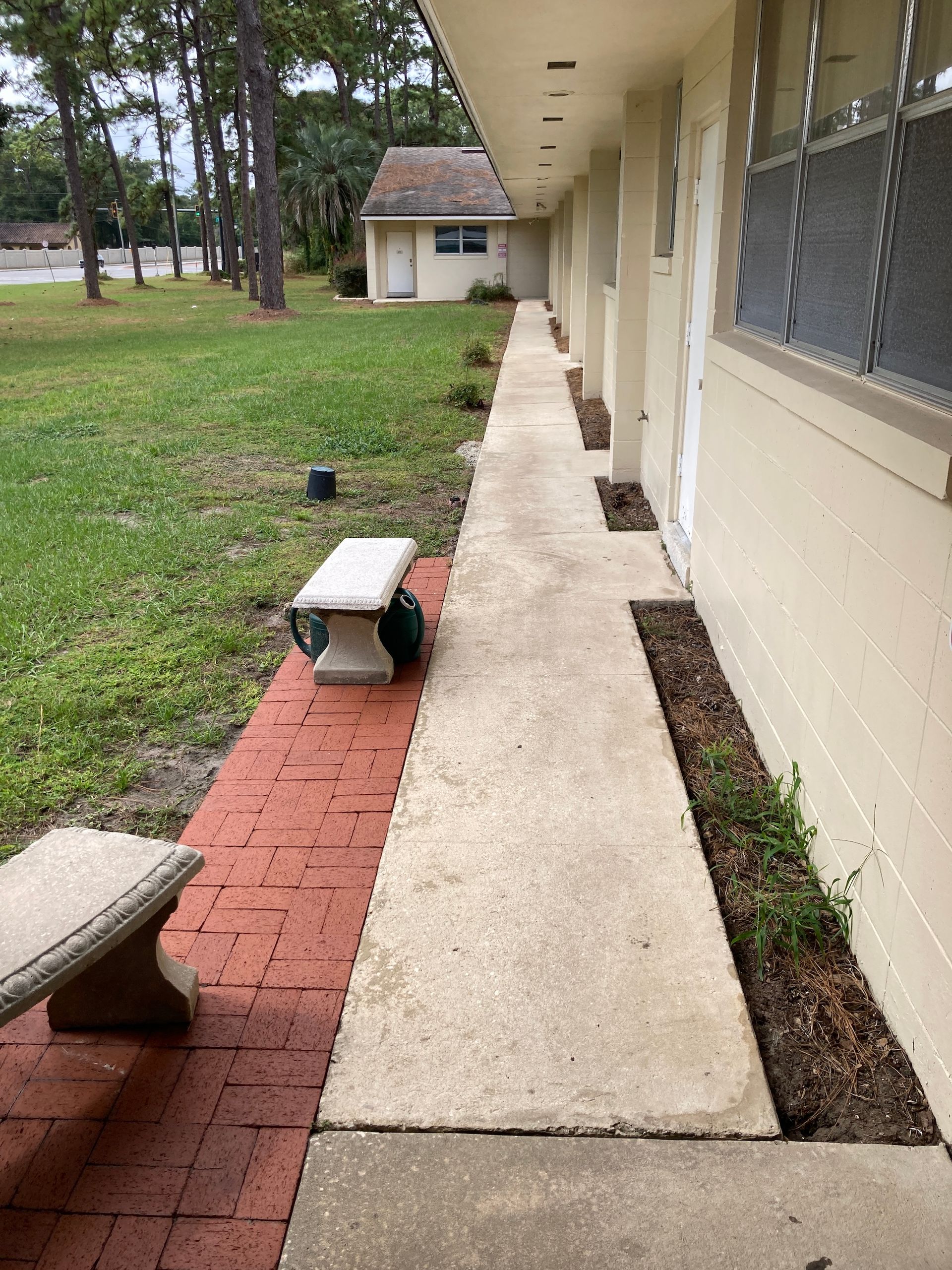 A concrete walkway alongside a building, bordered by a red brick path with two stone benches, leading to a small house.