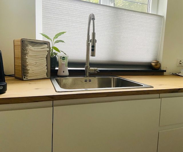 Moderne keuken met wastafel, chromen kraan, houten aanrechtblad, potplant en raam met jaloezieën.