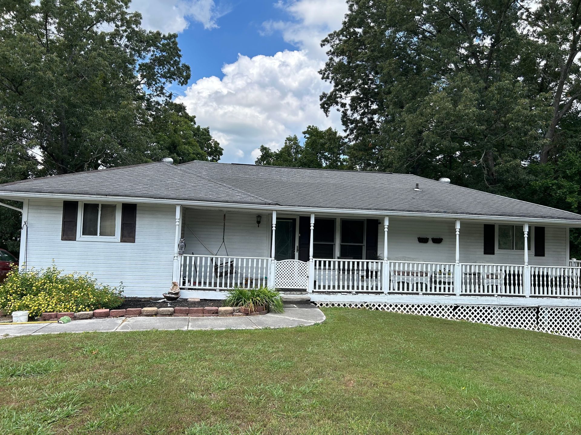 A white house with a gray roof and a large porch surrounded by trees.