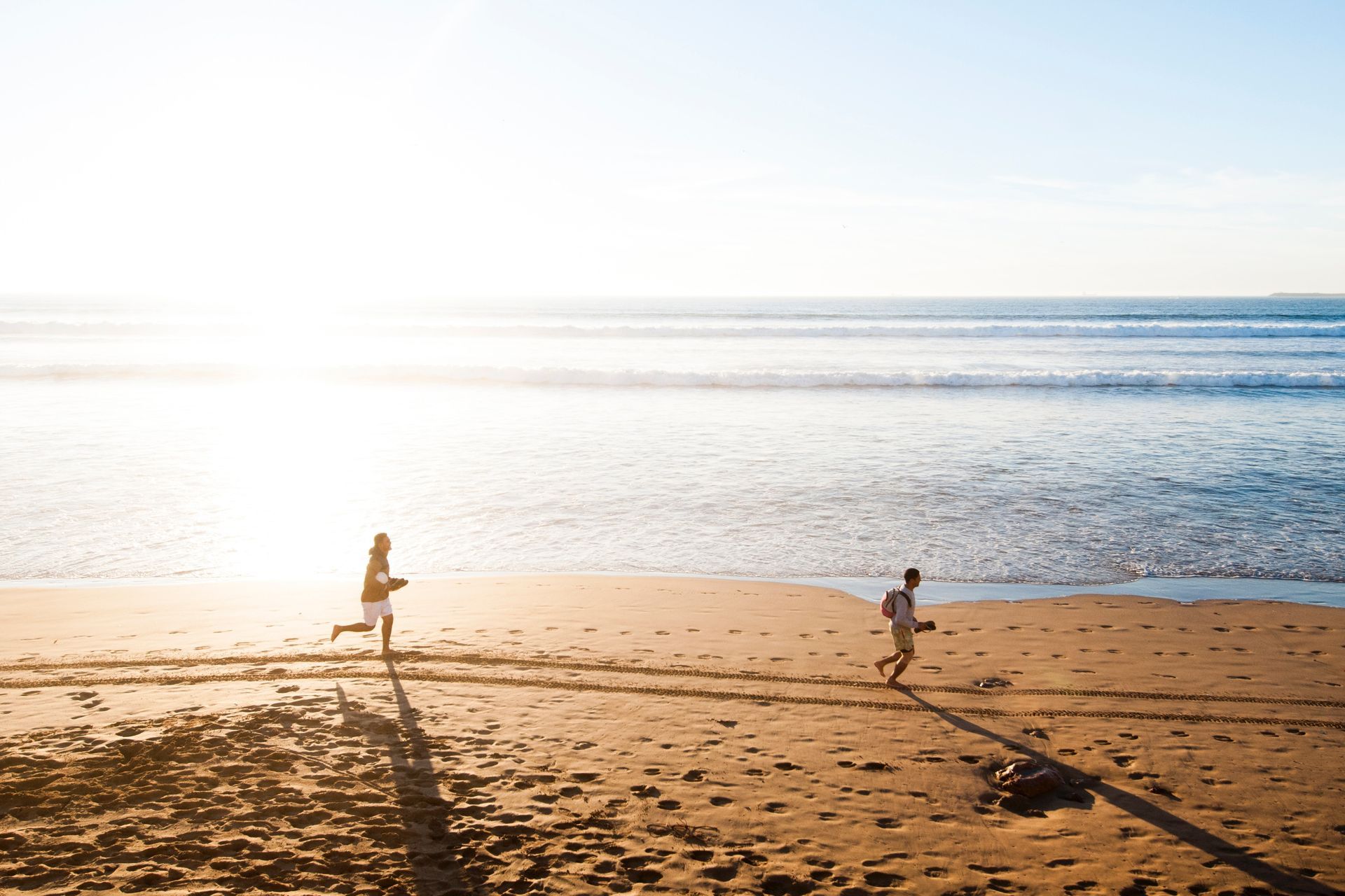 Two men running down a beach in the sand with the sun behind them.