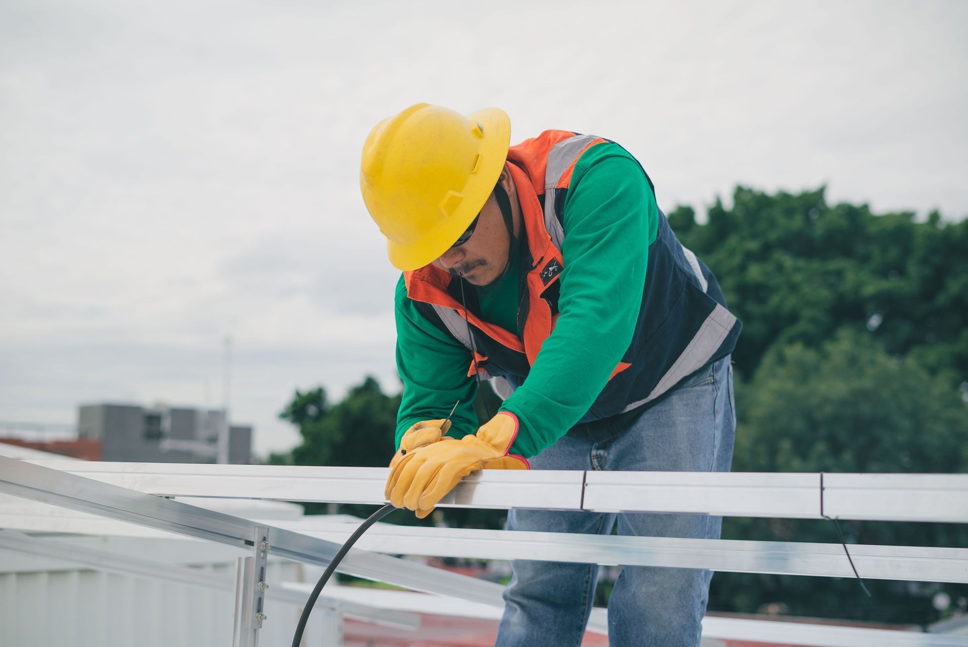 Worker in yellow hard hat and gloves securing rooftop metal framing outdoors.