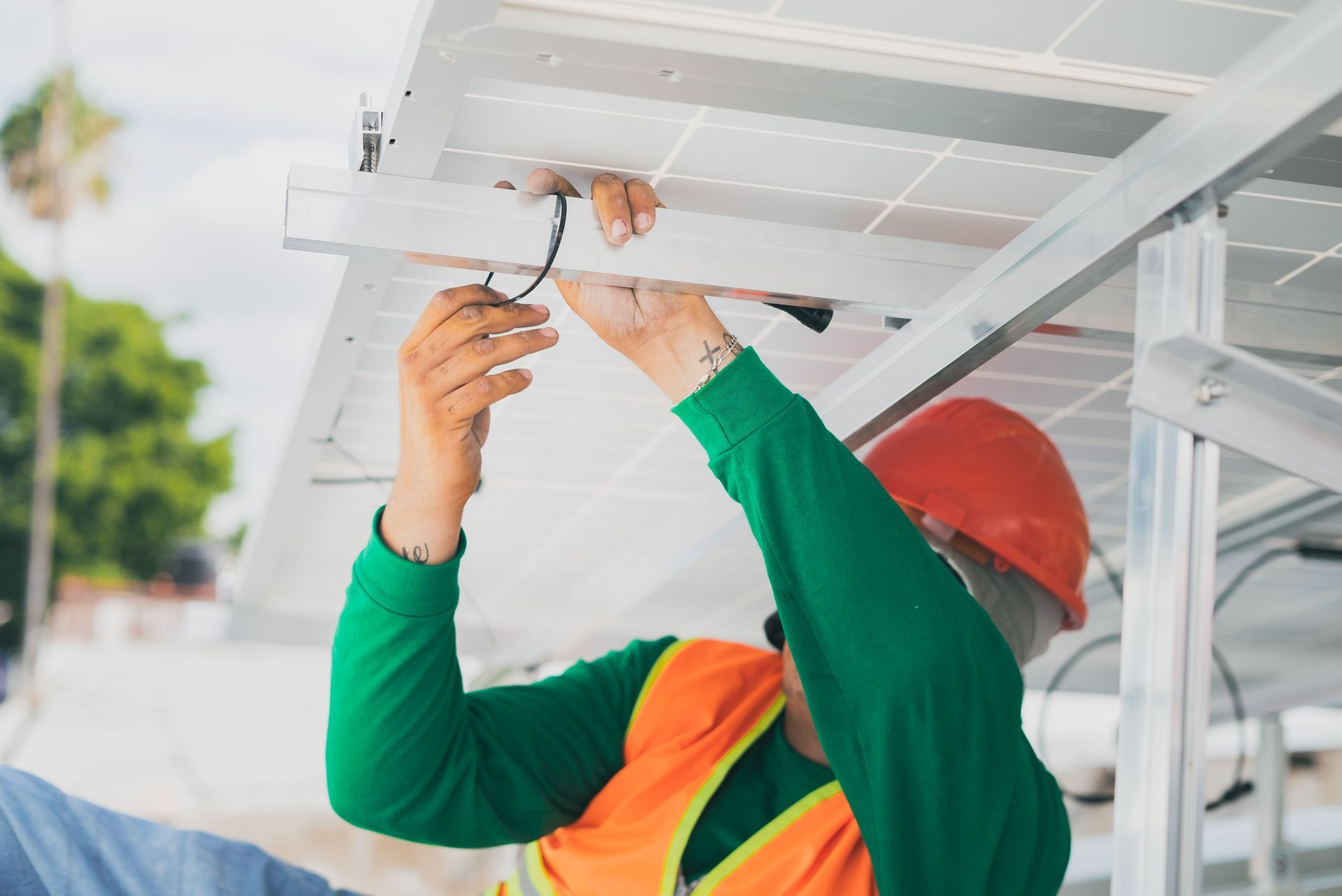 Worker in orange vest and red hard hat installing wiring under a solar panel array