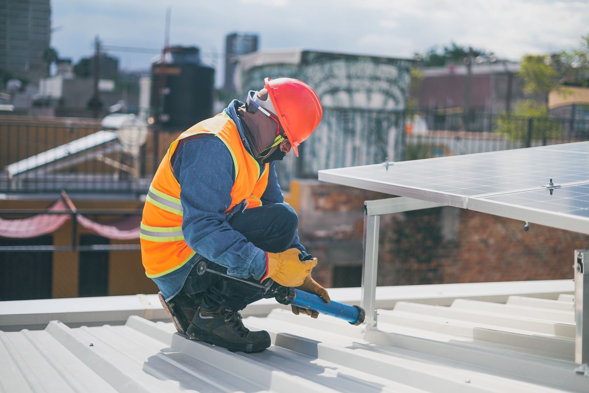 Worker in orange vest and hard hat installing rooftop solar panels on a white metal roof.