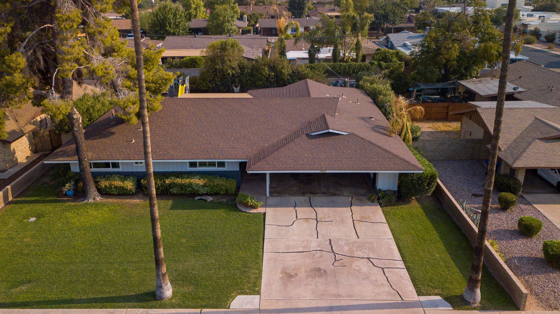 Aerial view of a single-story house with a driveway, green lawn, and palm trees in a suburban neighborhood