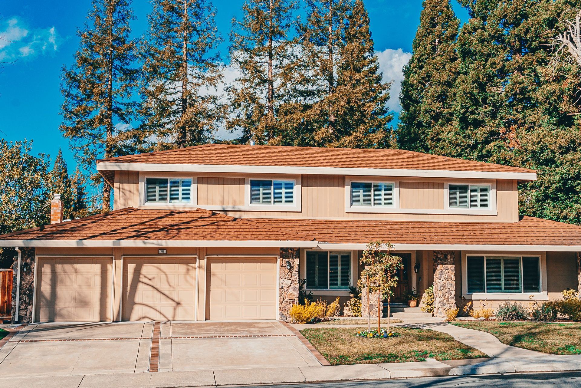 Two-story tan house with a garage, large windows, and tall trees under a bright blue sky