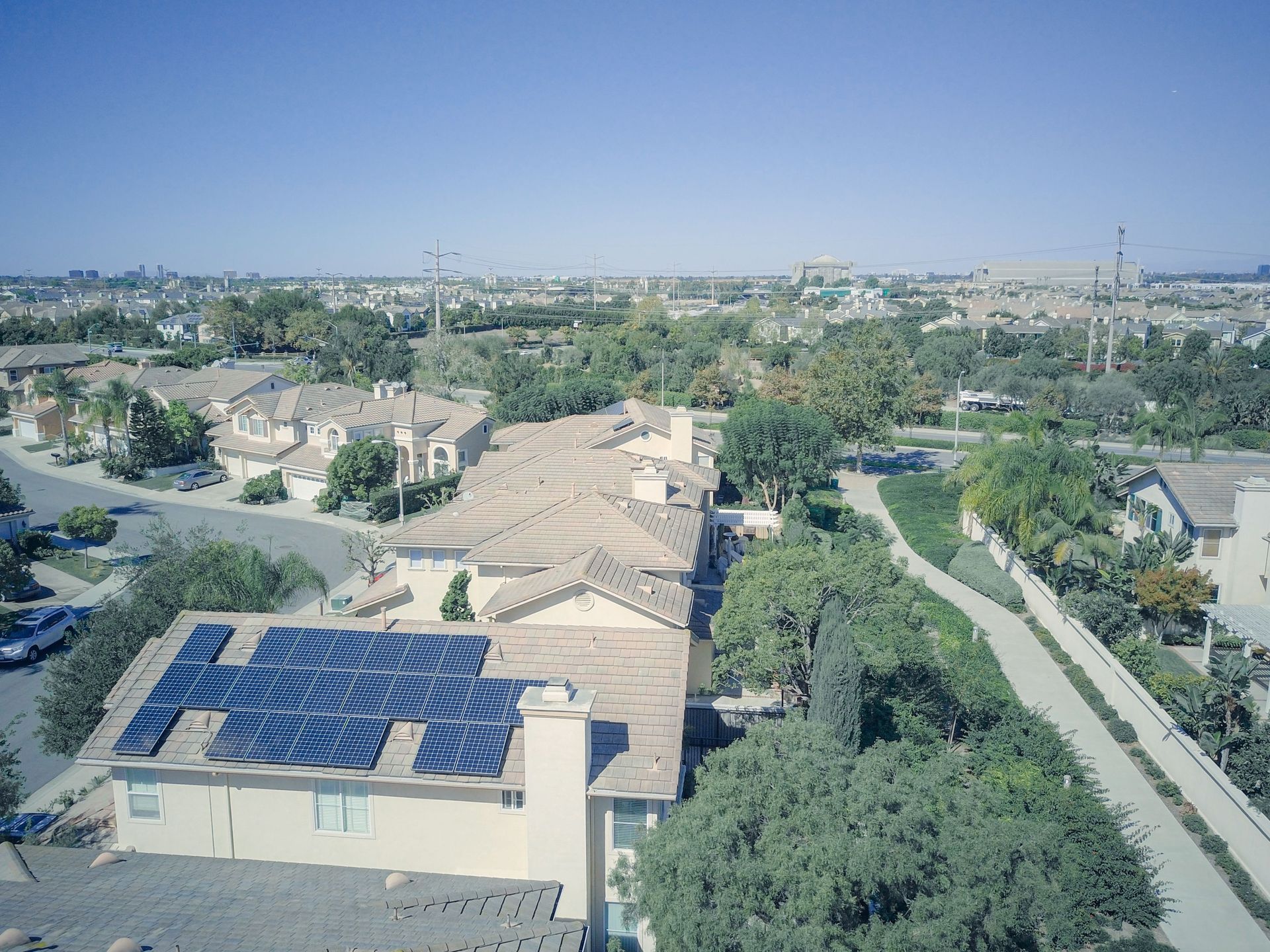 Aerial view of a suburban neighborhood with tan roofs, solar panels, trees, and a canal under a clear blue sky