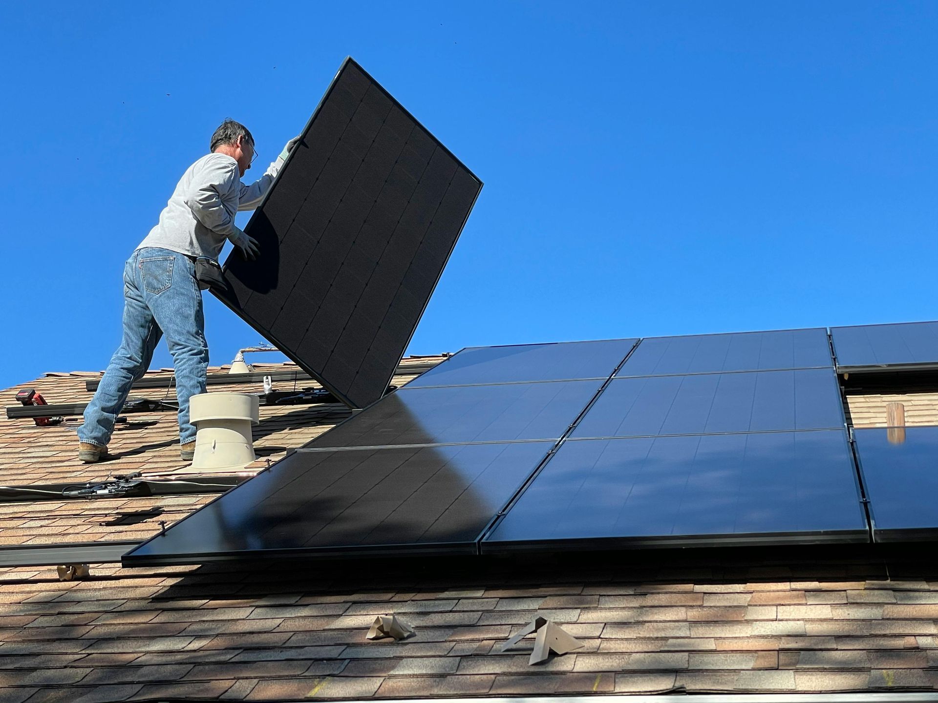 Worker installing solar panels on a shingled roof under a clear blue sky