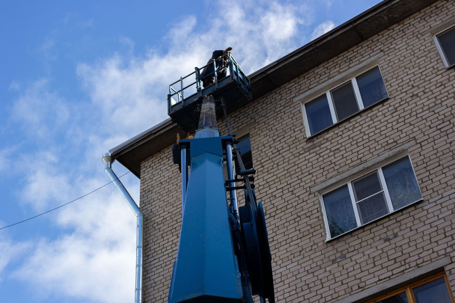 Blue cherry picker lift beside a brick building, reaching up to workers near the roofline under a cloudy sky