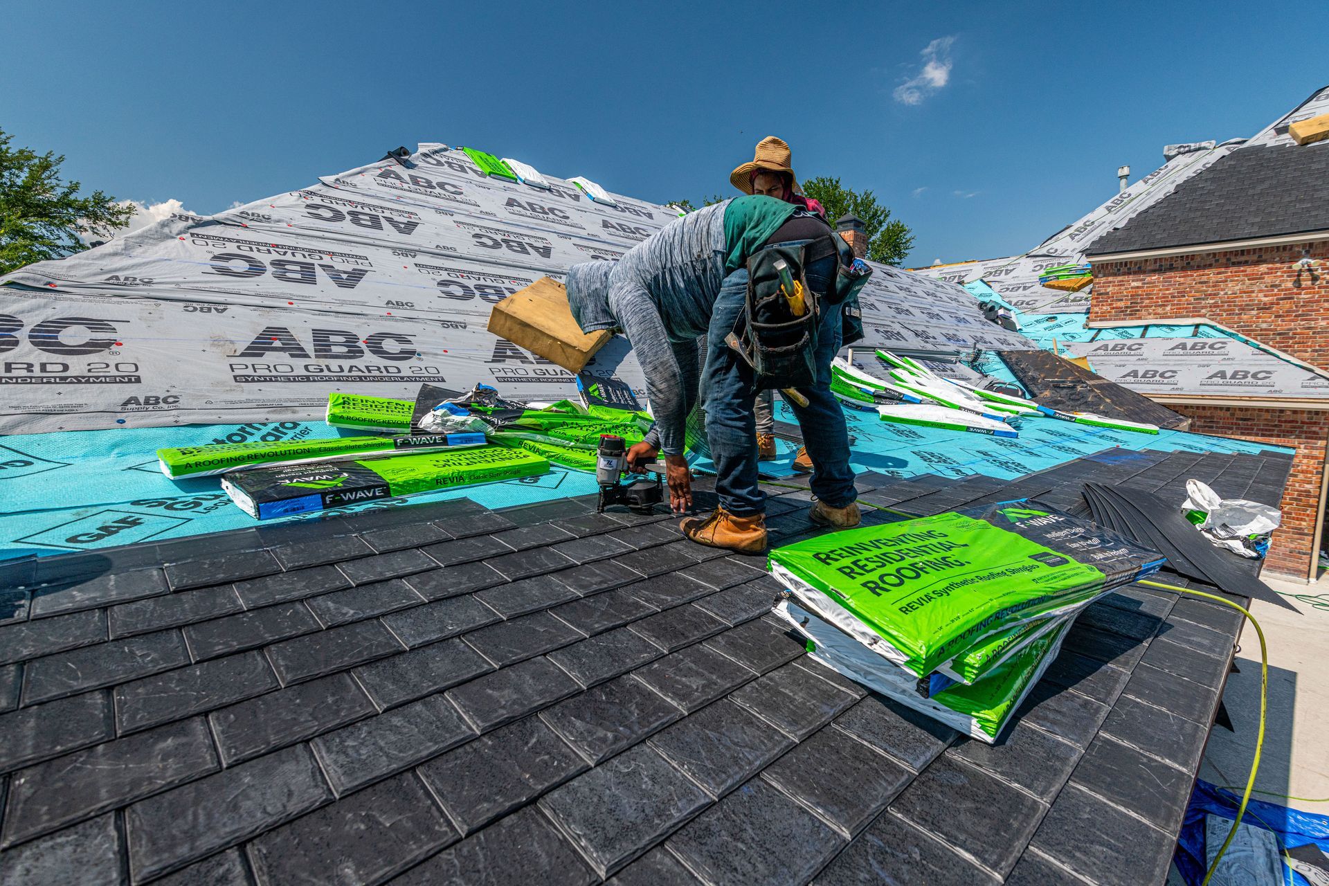 Workers on a roof installing bright green underlayment and black shingles under a blue sky