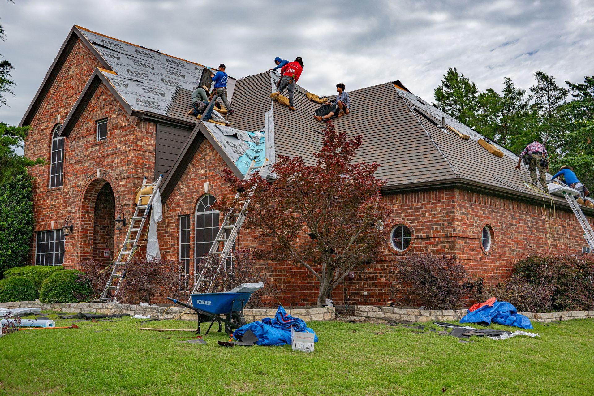 Brick house with damaged roof; workers are repairing it with blue tarps and debris in the yard