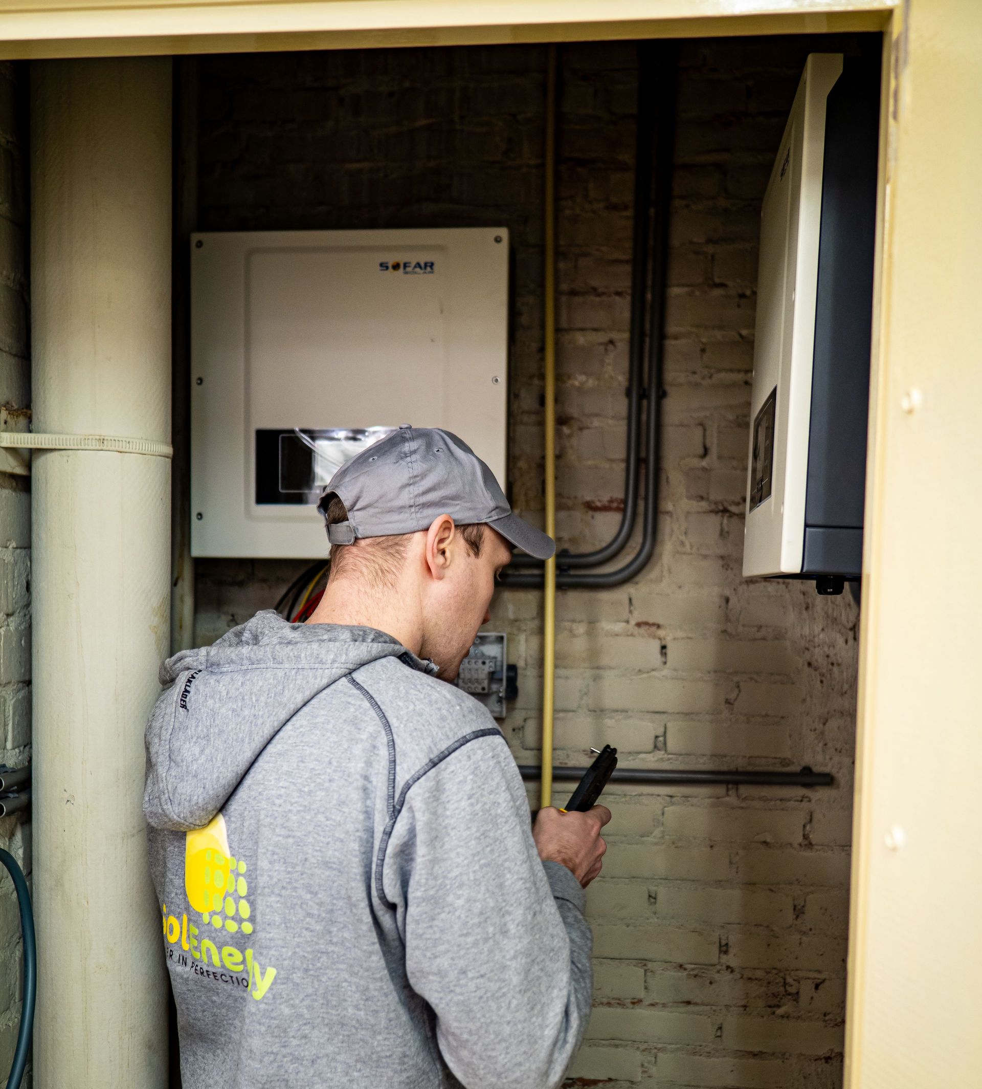 Person in gray hoodie and cap using a phone in a utility room with electrical panels and pipes