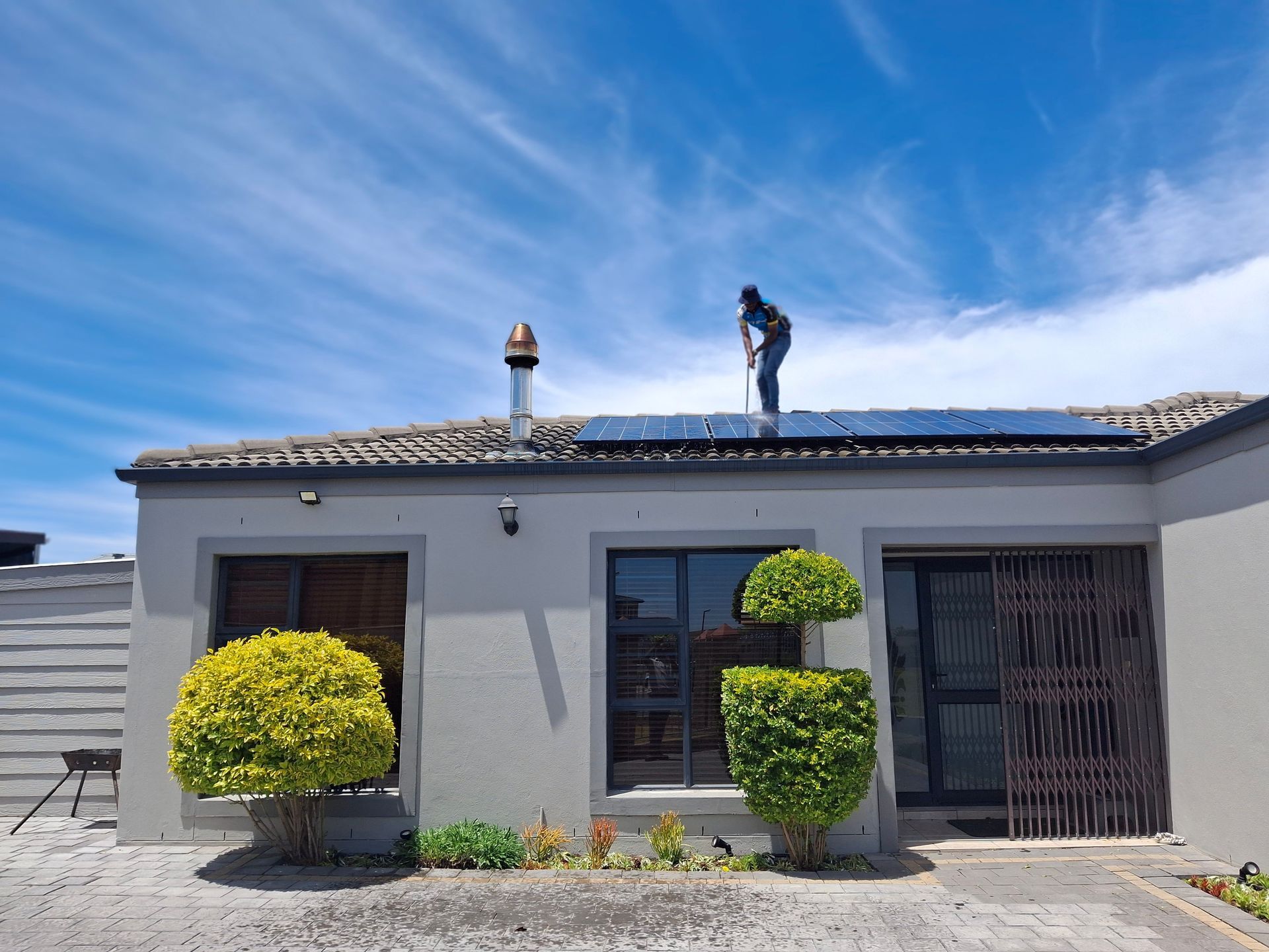 Roofer standing on a house roof with solar panels under a blue sky, trimmed shrubs below.
