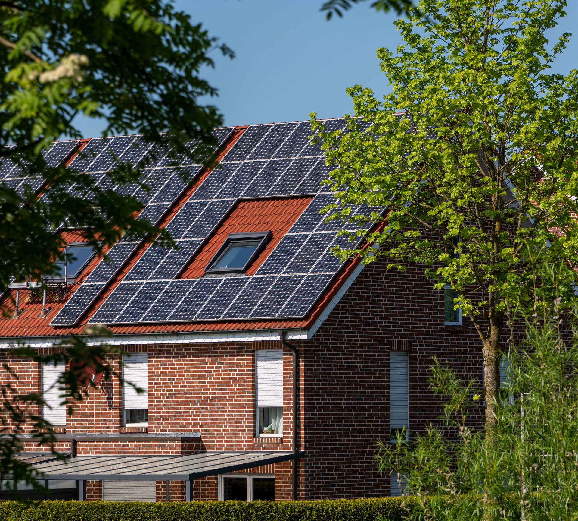 Brick house with solar panels on the roof, partly framed by green trees under a blue sky
