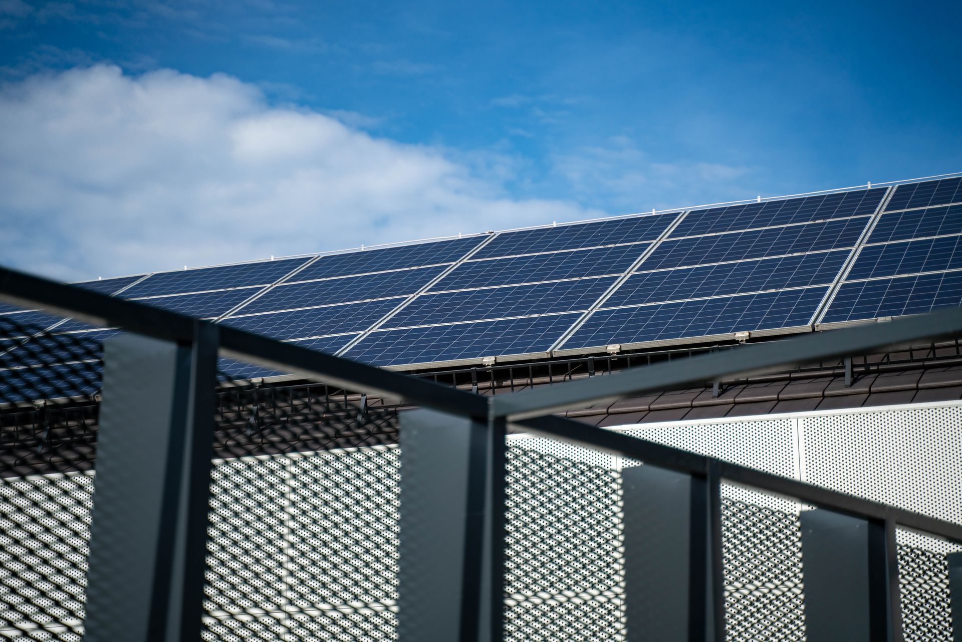 Solar panels mounted on a rooftop above a metal fence under a partly cloudy sky