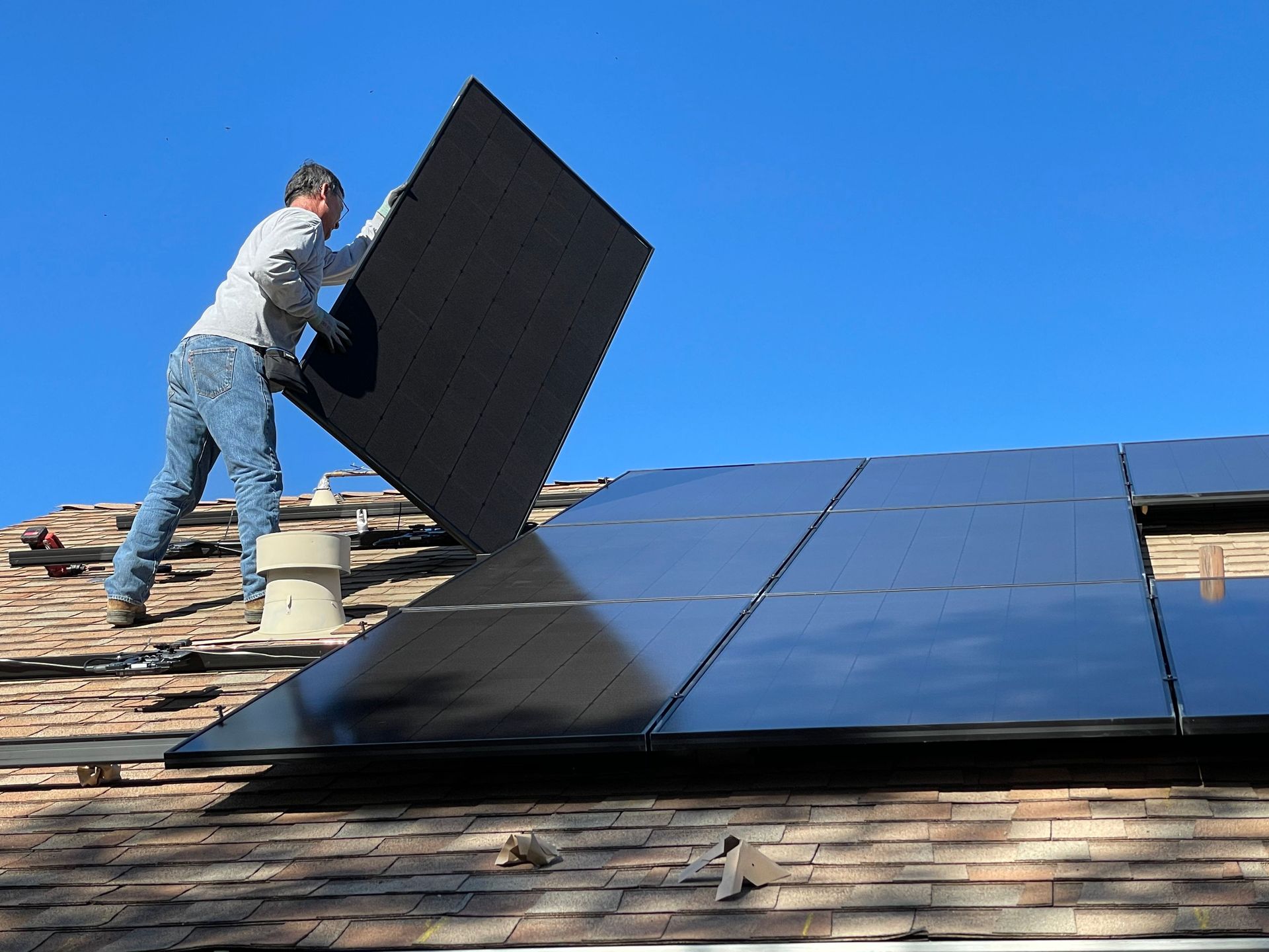 Worker installing black solar panels on a shingled rooftop under a clear blue sky