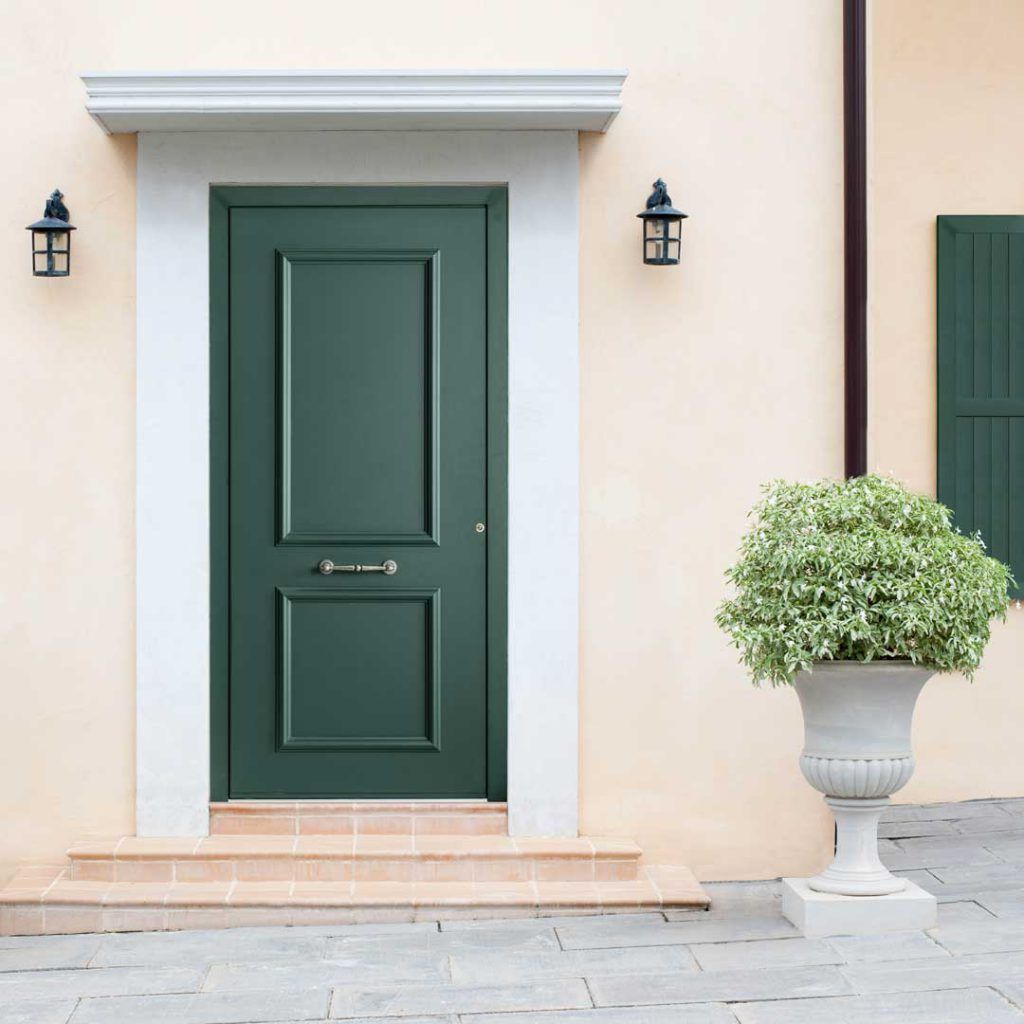 Porta d’ingresso con pannello verde, facciata beige, lanterne a parete e un arbusto in vaso sulla destra.