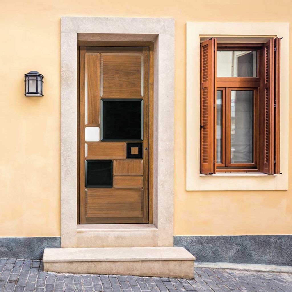 Porta d’ingresso e finestra in legno su una parete in stucco beige, con cornici in pietra e una piccola lanterna da parete.