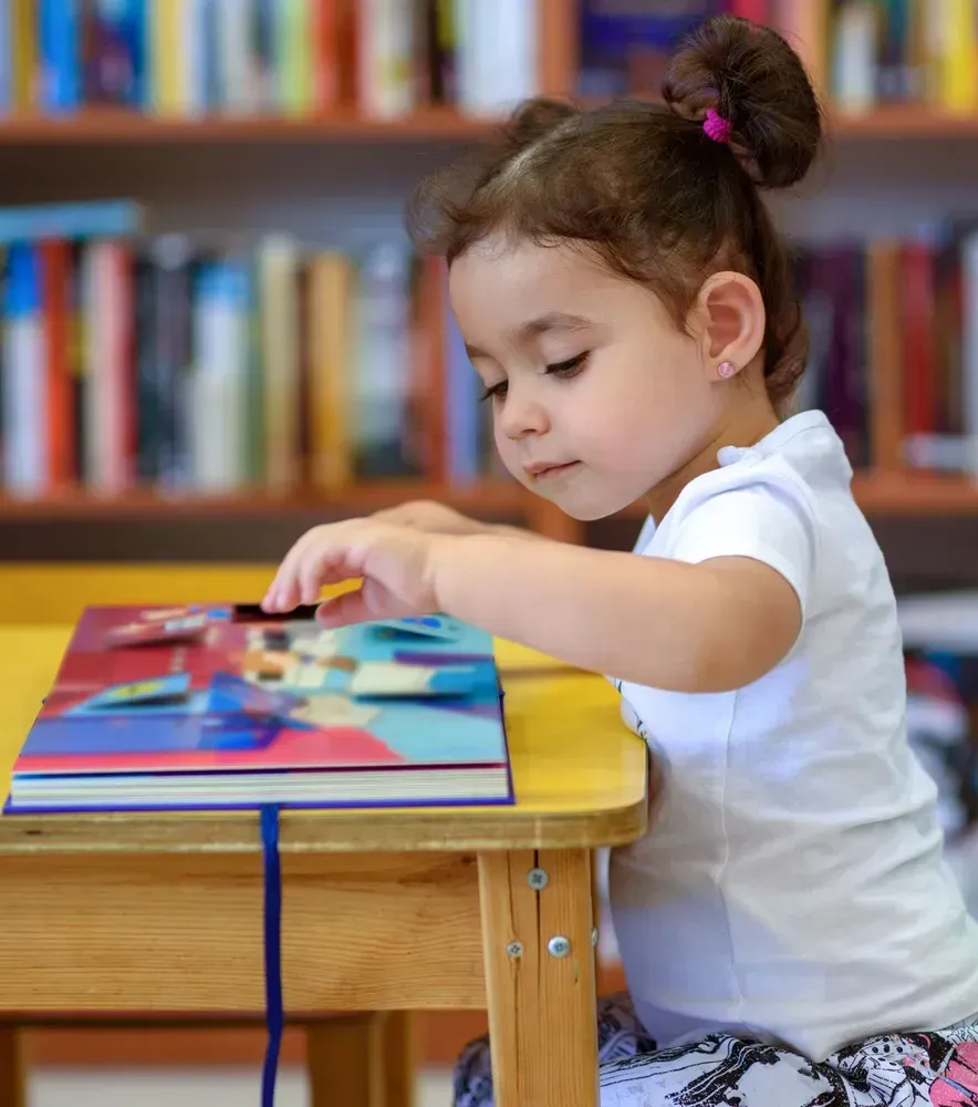 Child reading a colorful book at a wooden table in a library
