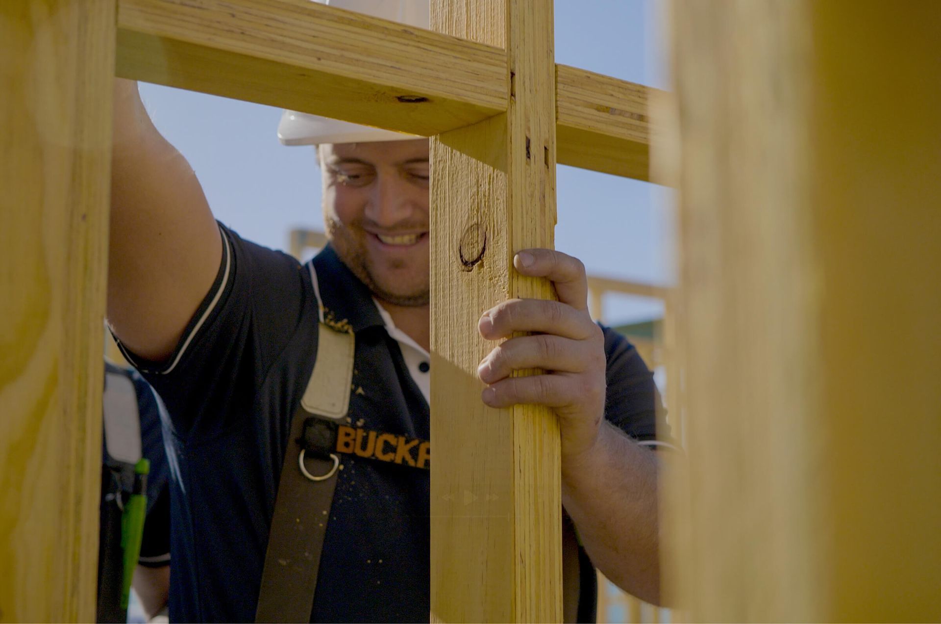 Construction worker smiling while assembling wooden frame. Outdoors, sunny.