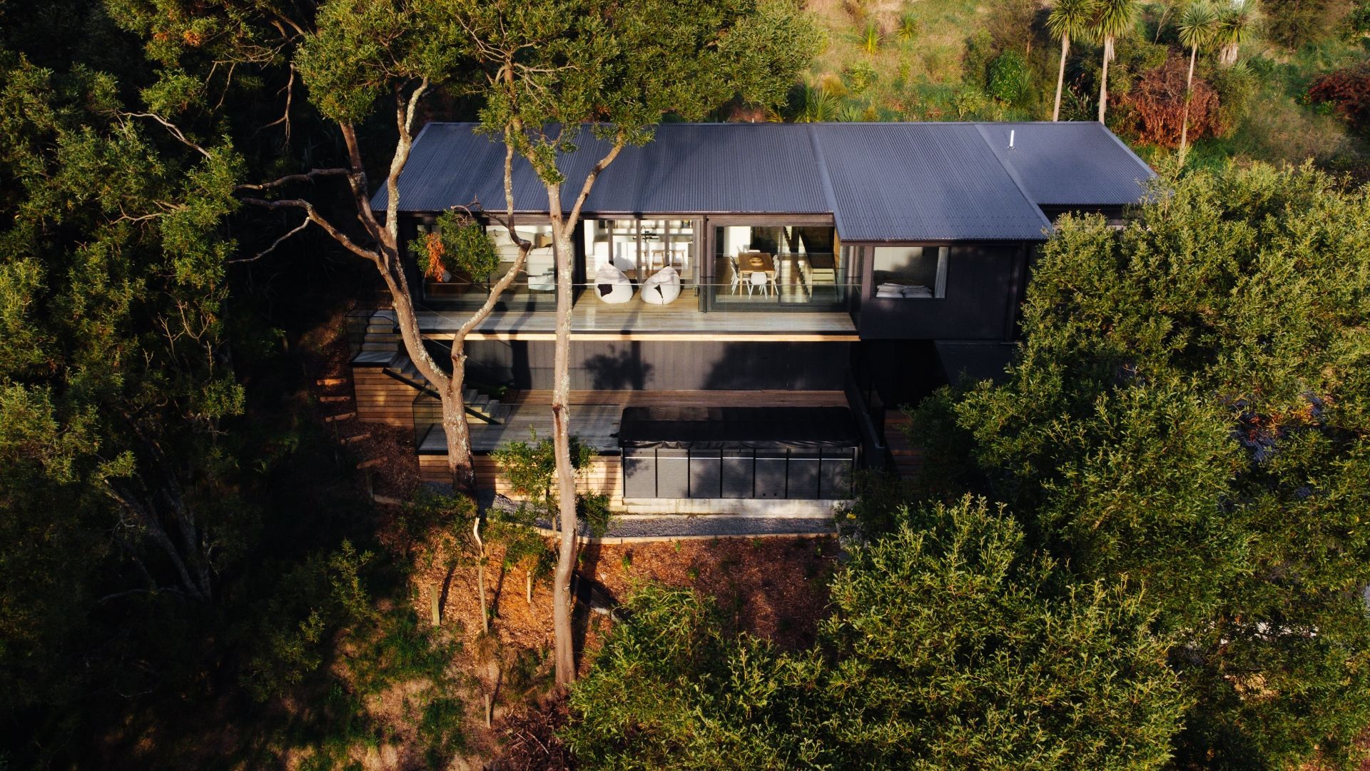 Modern, dark-colored house nestled among trees; glass walls, deck, and black roof.