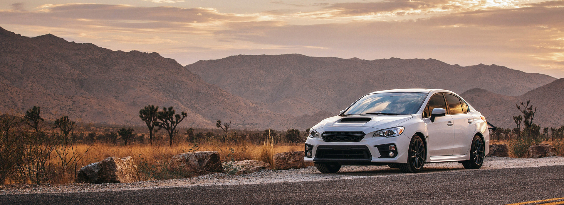White Subaru WRX sedan parked on a road in a desert landscape with mountains in the background at sunset. | King's Auto Center