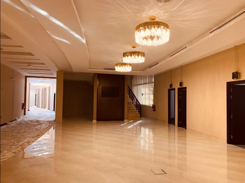 Brightly lit interior hallway with gold chandeliers, beige walls, and reflective marble flooring. Three black doors and a staircase are visible.