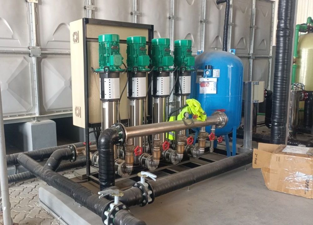 Water pump system with four vertical green pumps, a blue pressure tank, and black pipes in an industrial setting.
