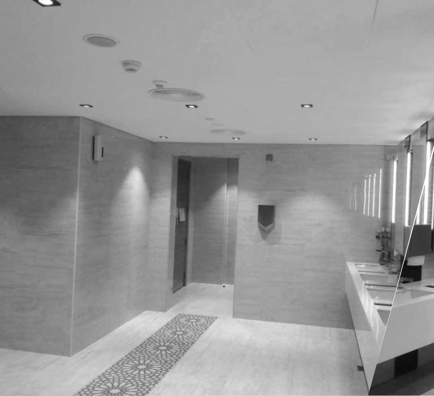 Black and white photograph of a modern restroom interior. Shows tiled walls and ceiling with recessed lighting, a doorway, and a counter.