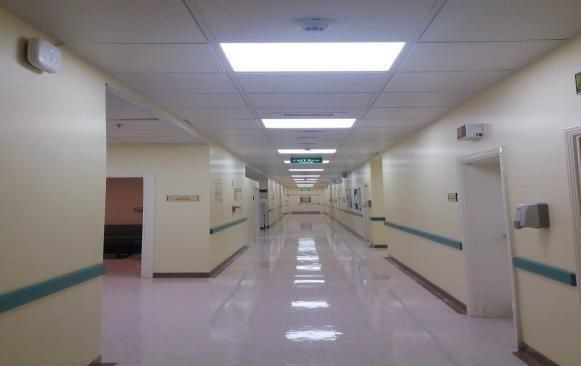 Hospital hallway with cream walls, blue handrails, and fluorescent lighting. Doors and signage line the sides, leading to a distant green exit sign.
