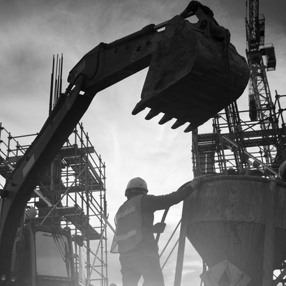 A construction worker in a hard hat and vest stands near an excavator, with scaffolding and a cloudy sky in the background.