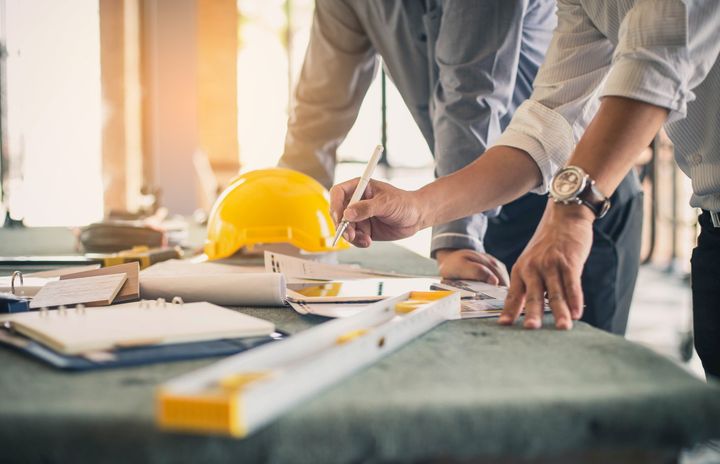 Two people reviewing blueprints on a table, with a yellow hard hat, tools, and a level visible.