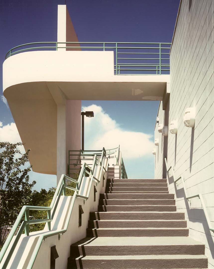 Exterior staircase leading to a white, curved platform with green railings. Blue sky with clouds.