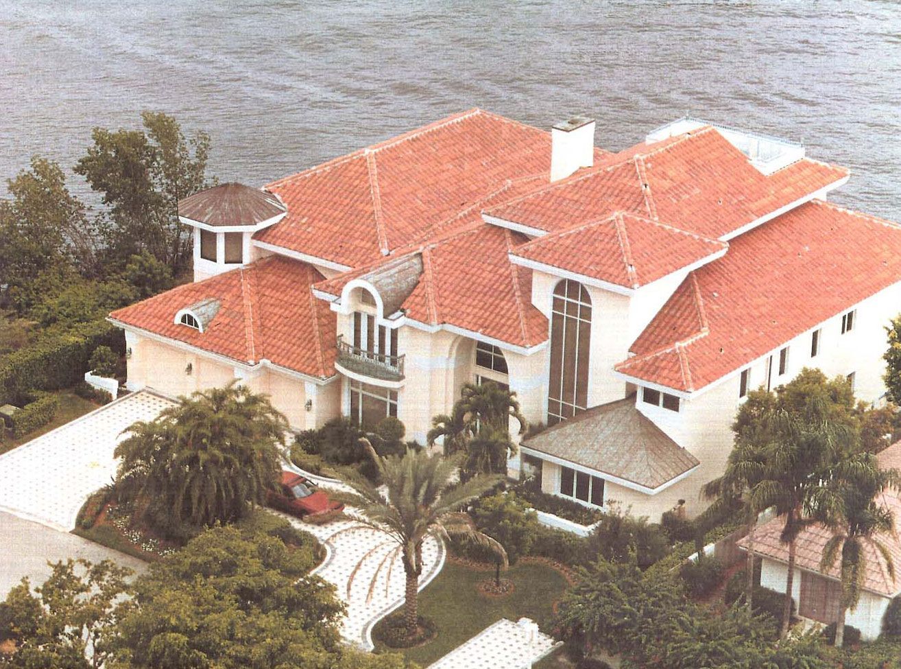 Luxury house with a red-tiled roof, near water, with a long driveway and tropical landscaping.