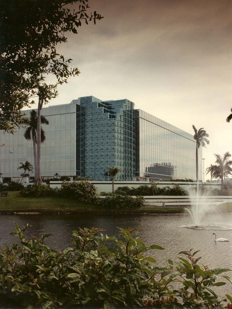 Glass-walled building near a lake with fountain and palm trees under an overcast sky.