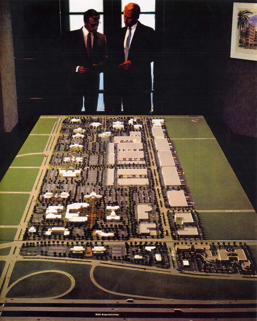 Two men in suits look at a model of a large planned development with buildings and roads.