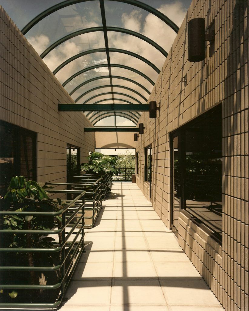 A covered walkway with a glass arched roof and tiled floor. Walls are tan brick with windows and railings.