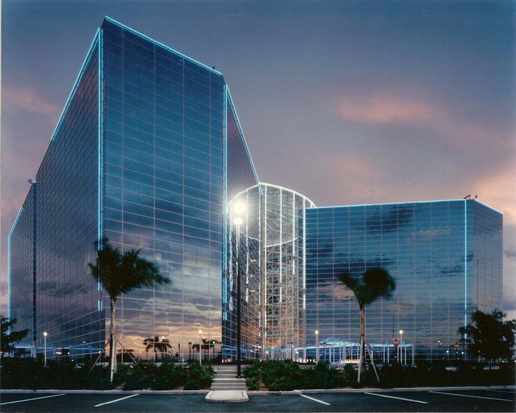 Modern glass building with blue-lit edges, palm trees in foreground, and sunset sky.