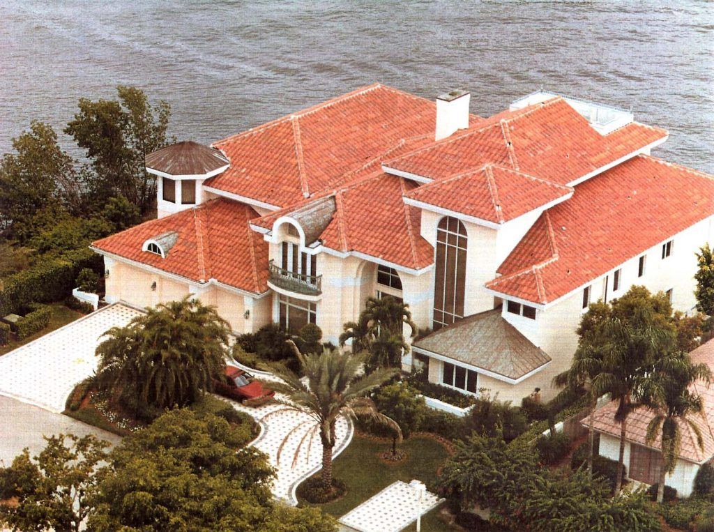 Large house with a red-tiled roof, waterfront view, and palm trees in the yard.