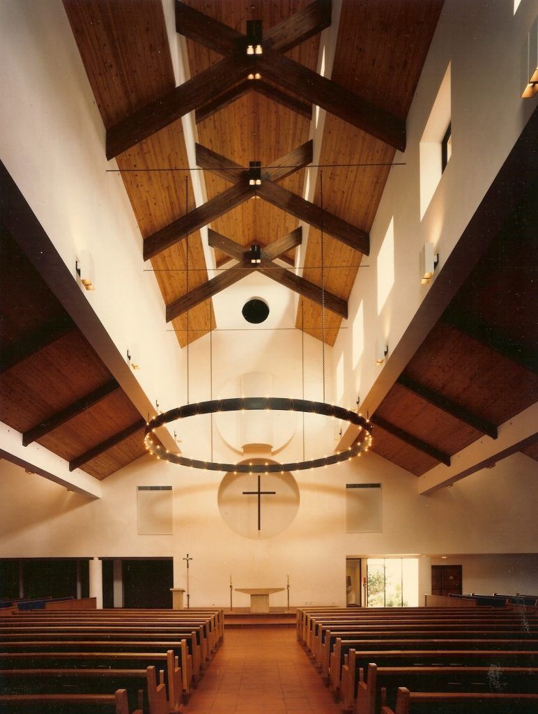 Interior of a church with wooden beams on the ceiling, rows of pews, and a cross on the wall.