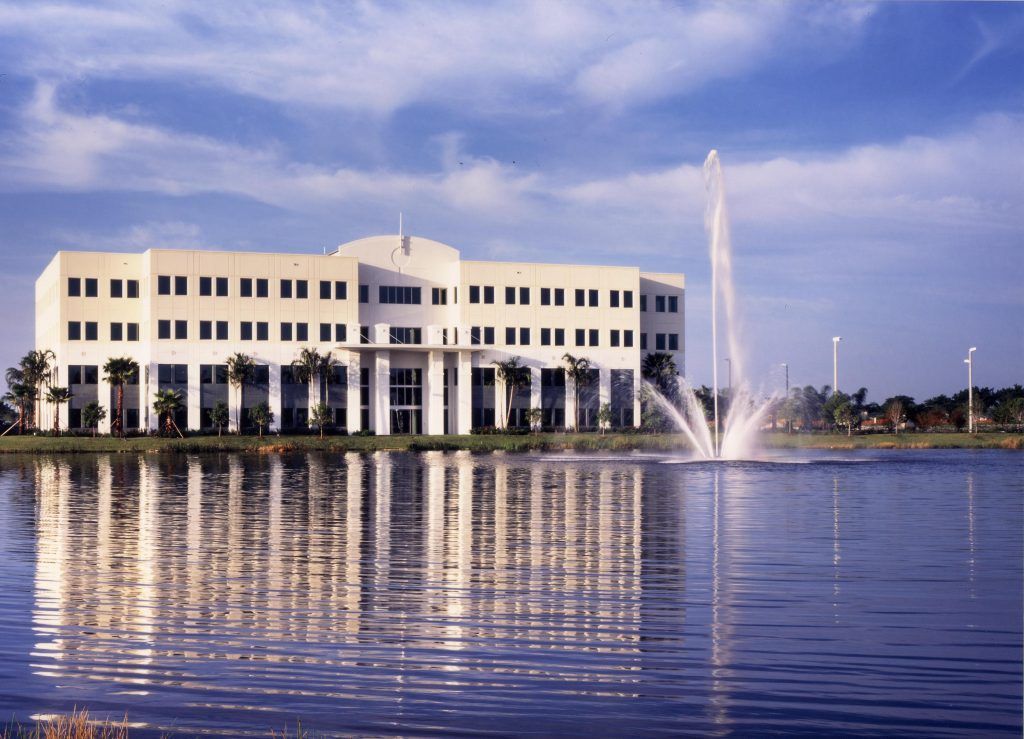 Office building reflected in lake, fountain sprays, sunny day, blue sky.
