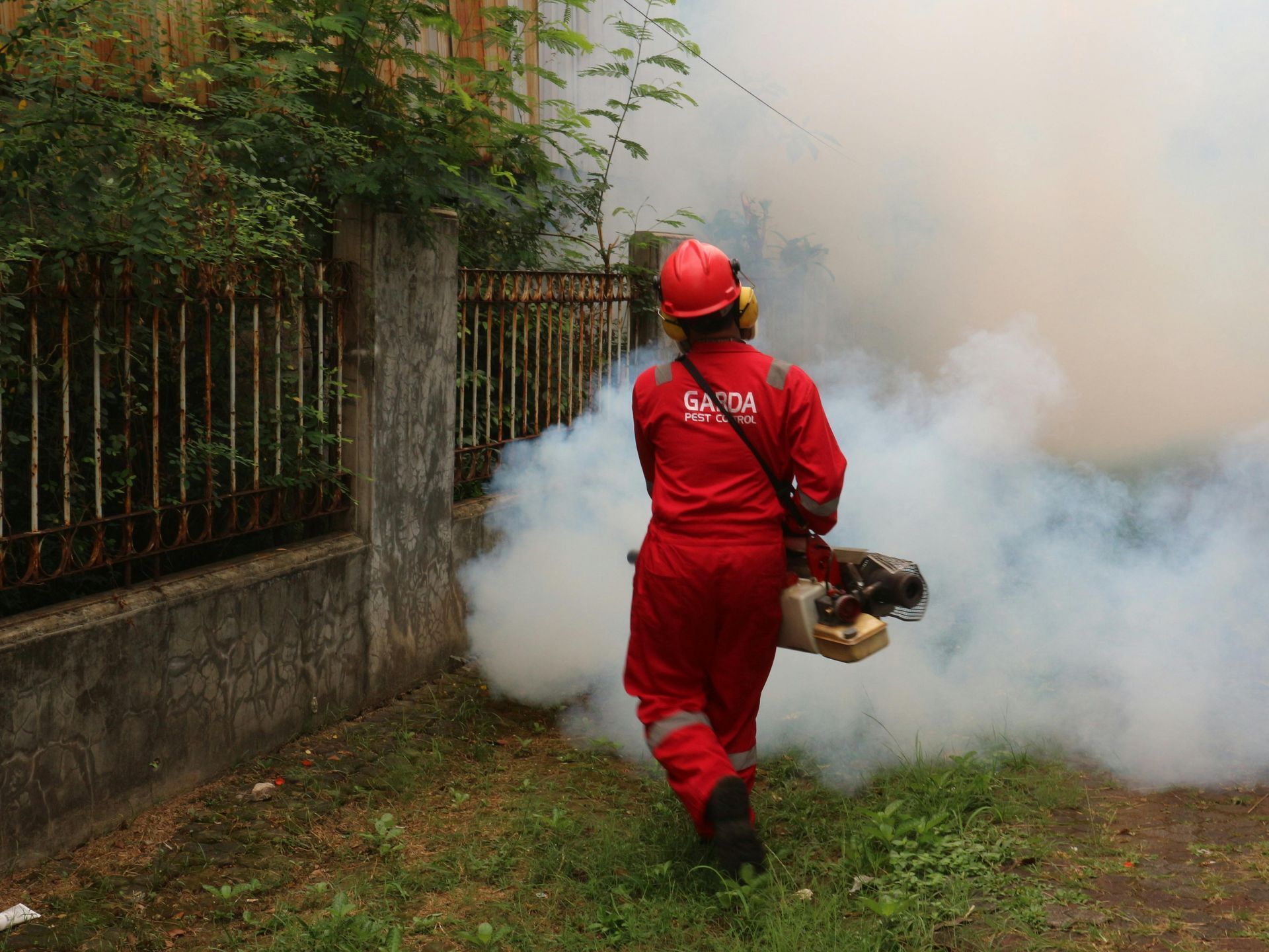 Firefighter in red gear carries a smoking object through thick smoke beside a fence.