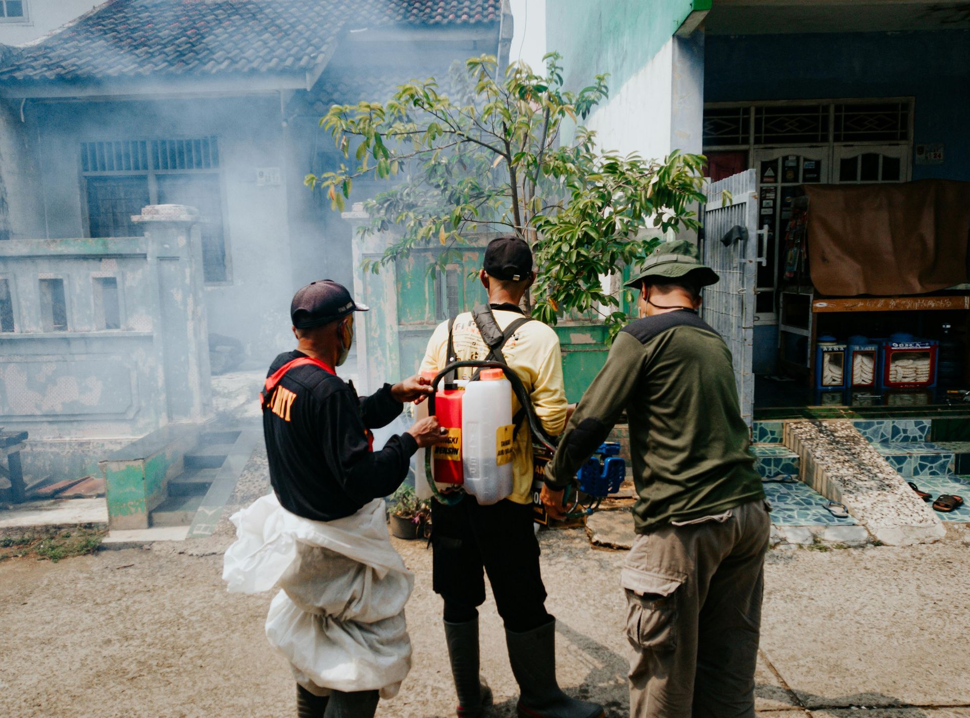 Three workers spray insecticide near a house, one wearing a backpack sprayer amid smoke.