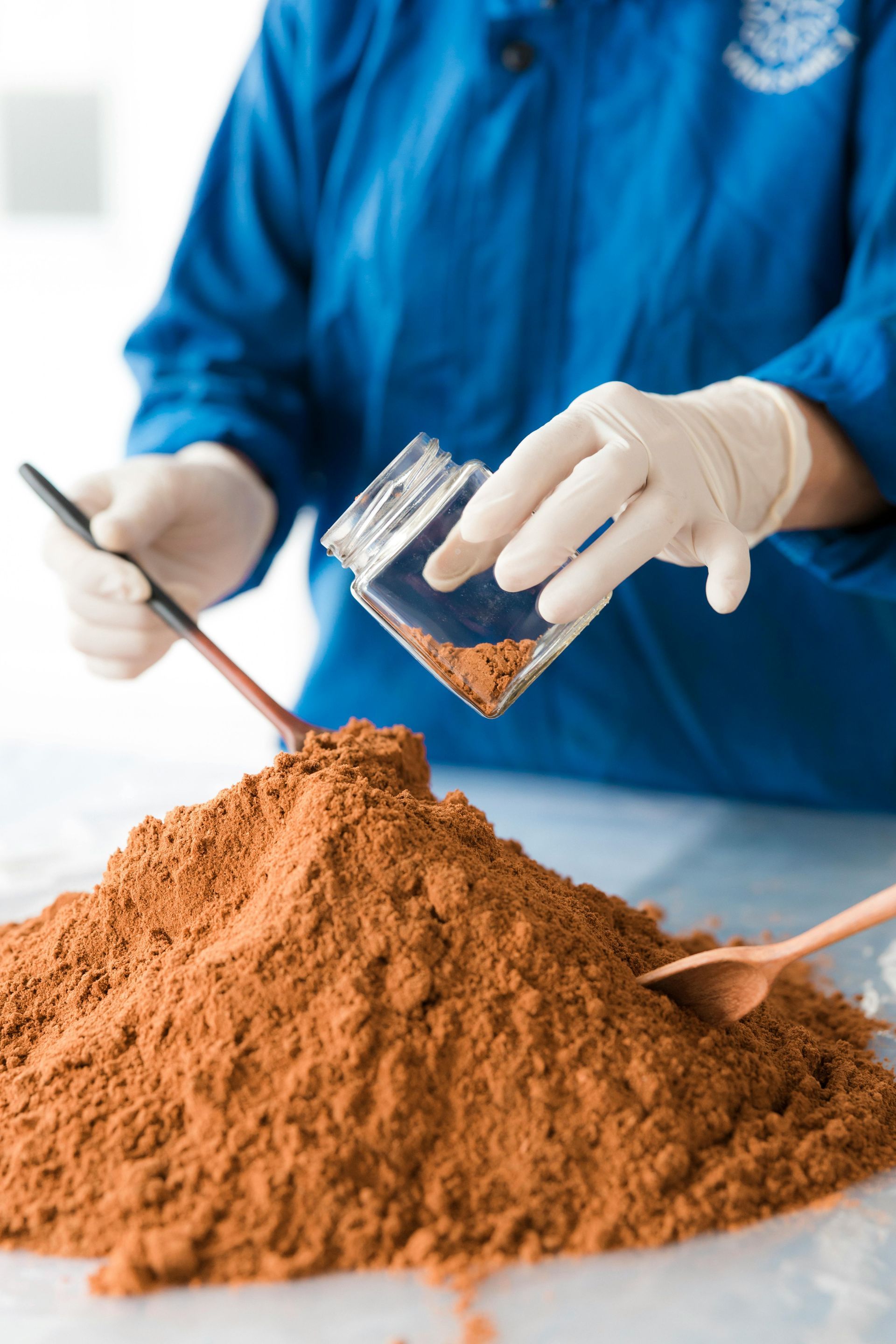 Gloved hands sifting spice powder in a bowl, with a small jar and spoon on the table