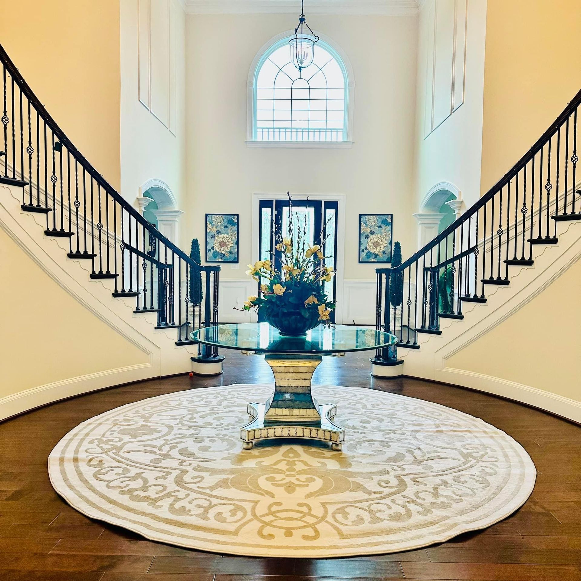 Grand staircase foyer with black railings, central floral arrangement, and a round patterned rug under a tall arched window