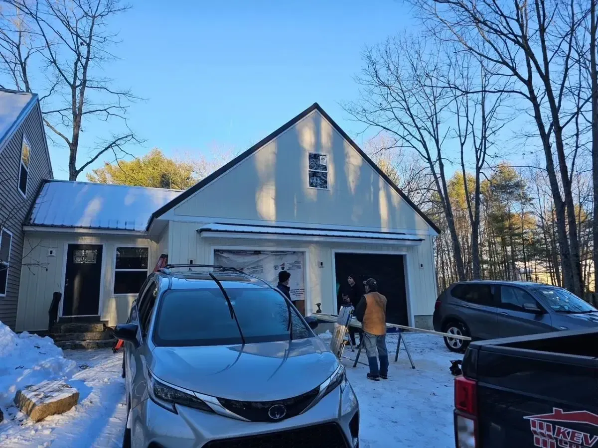 Exterior of a white house with black trim. Snow covers the ground. People work outside near a garage with two cars parked in front.