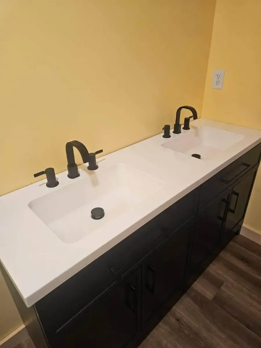 Double vanity bathroom sink with white countertop, black faucets, and dark cabinets against a yellow wall